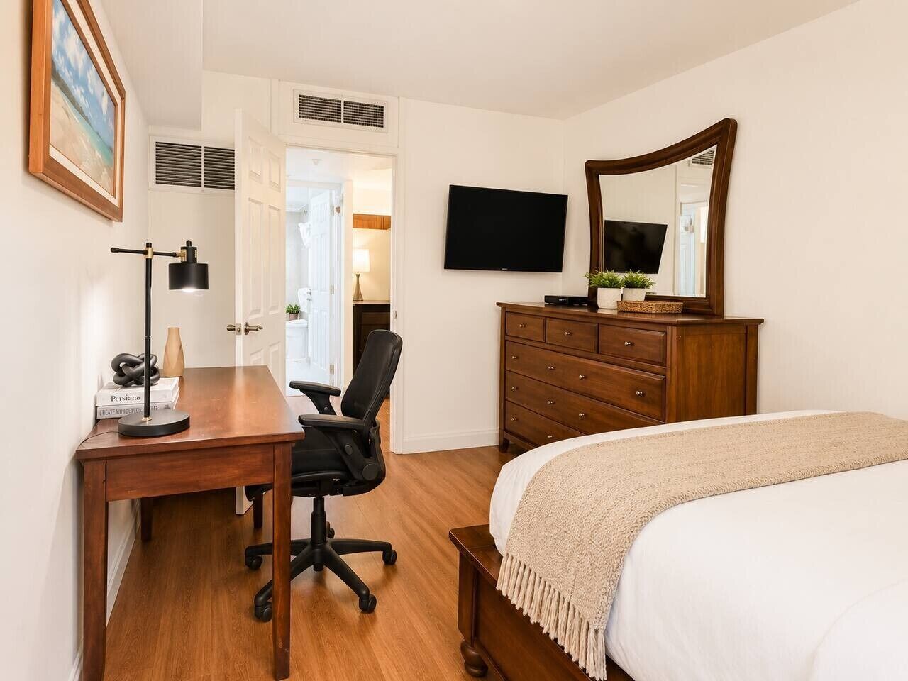 A hotel room with a wooden desk, black office chair, a bed with a white blanket, a wooden dresser, a flat-screen TV, and a large mirror.