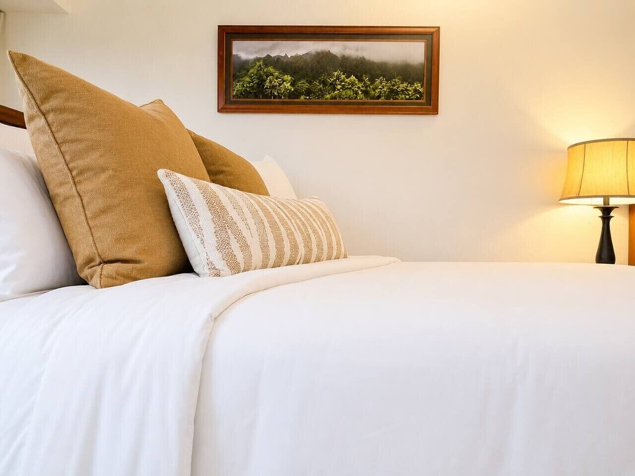 Un lit bien fait, avec une literie blanche et deux oreillers, un tableau encadré représentant un paysage accroché au mur, et une lampe sur la table de chevet.