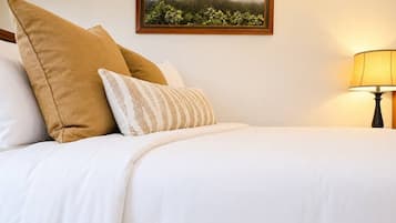 A neatly made bed with white bedding and two pillows, a framed picture of a landscape on the wall, and a lamp on the side.