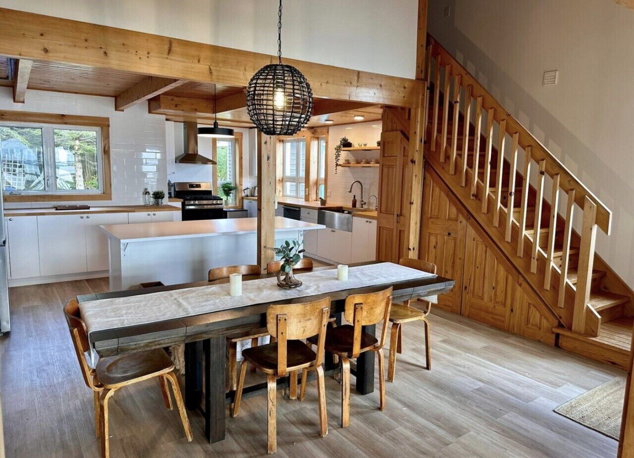 Una cocina espaciosa con vigas de madera, un comedor con una mesa larga y sillas, y una escalera con barandillas de madera.