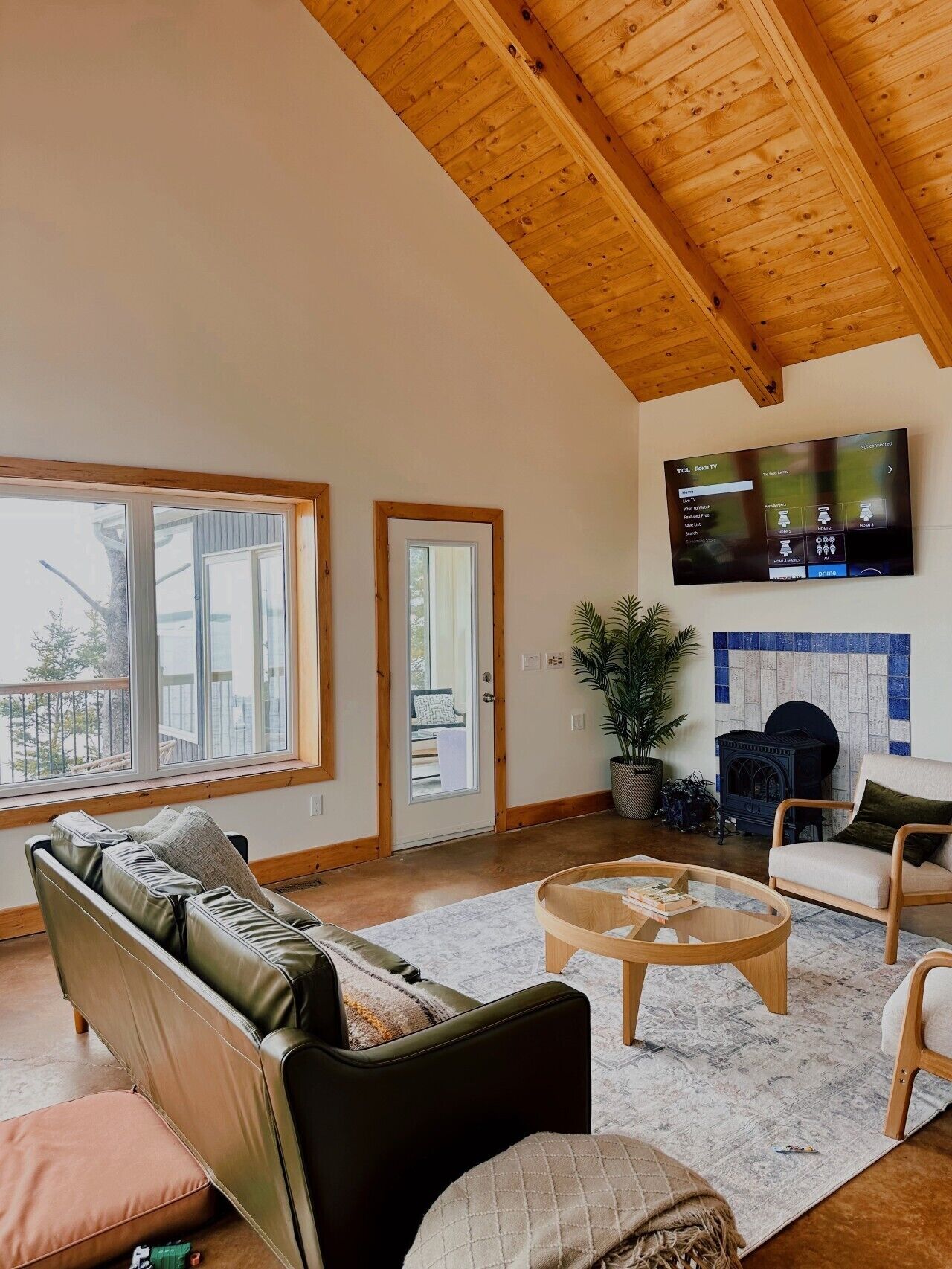 A spacious living room with a high ceiling, a large flat-screen TV mounted on the wall, a leather sofa, a wooden coffee table, and a potted plant.