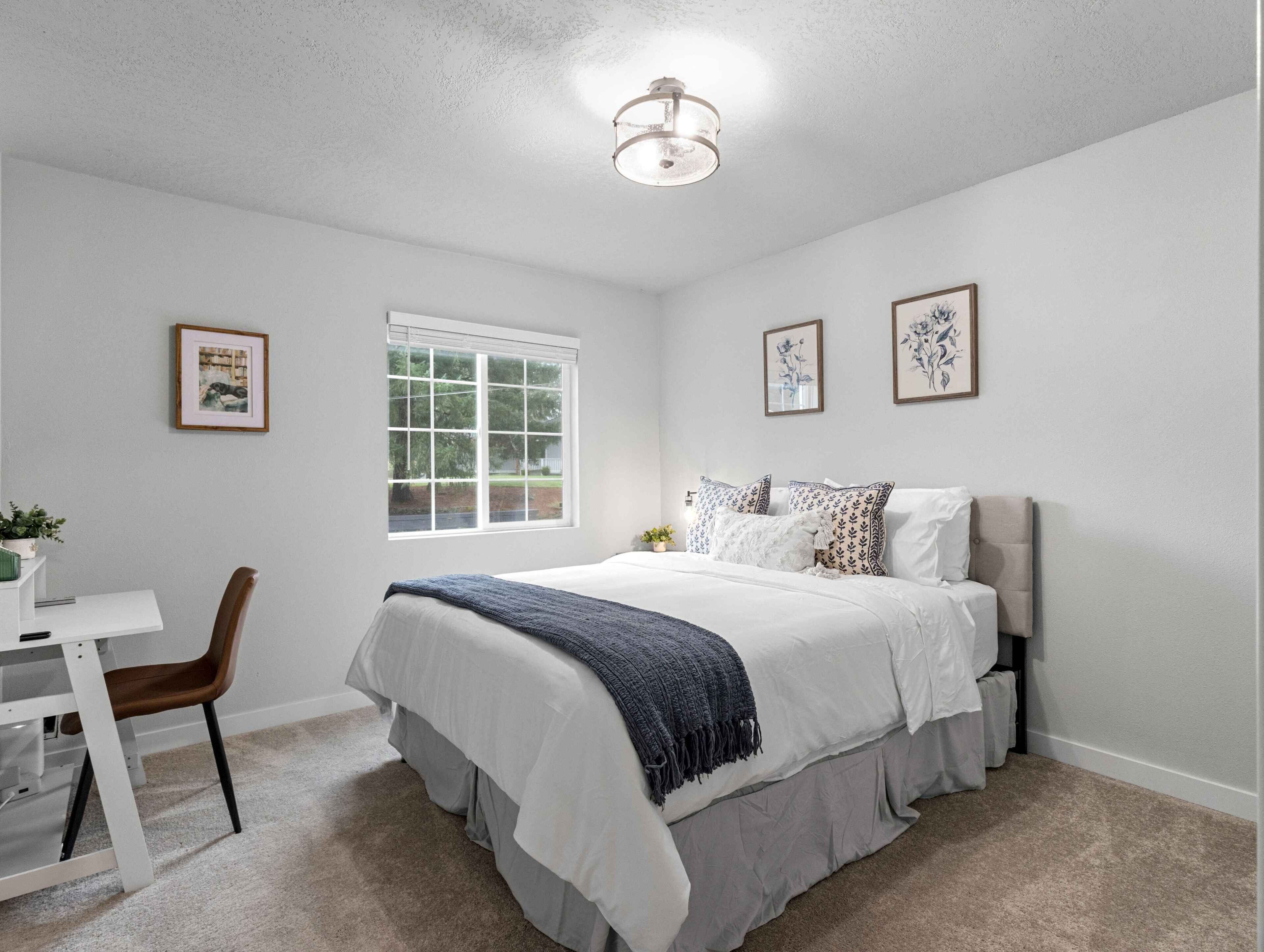 A bedroom with a bed, a desk, a chair, a window, and framed pictures on the wall.