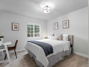 A bedroom with a bed, a desk, a chair, a window, and framed pictures on the wall.