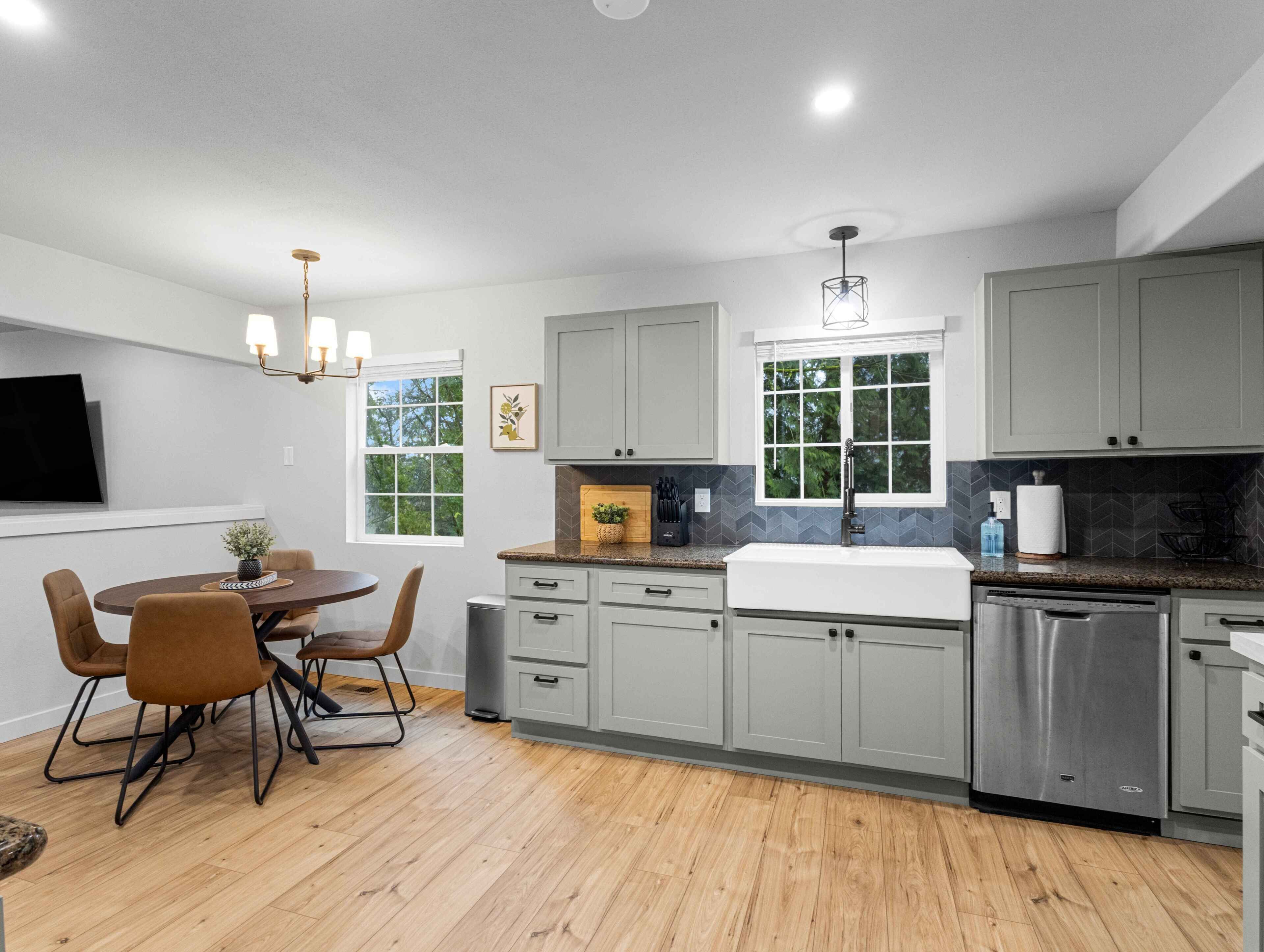 A modern kitchen with grey cabinets, a stainless steel dishwasher, and a dining area with a round table and chairs.