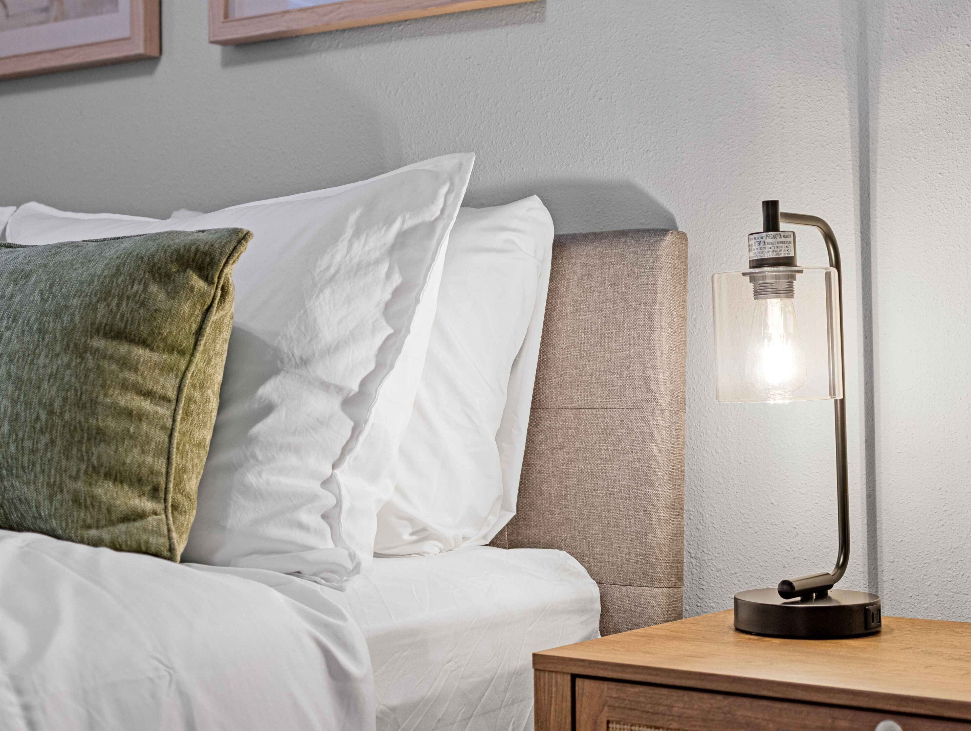 Uma cama bem arrumada com almofadas brancas e verdes, uma mesinha de cabeceira de madeira com uma luminária moderna e uma parede texturizada ao fundo.