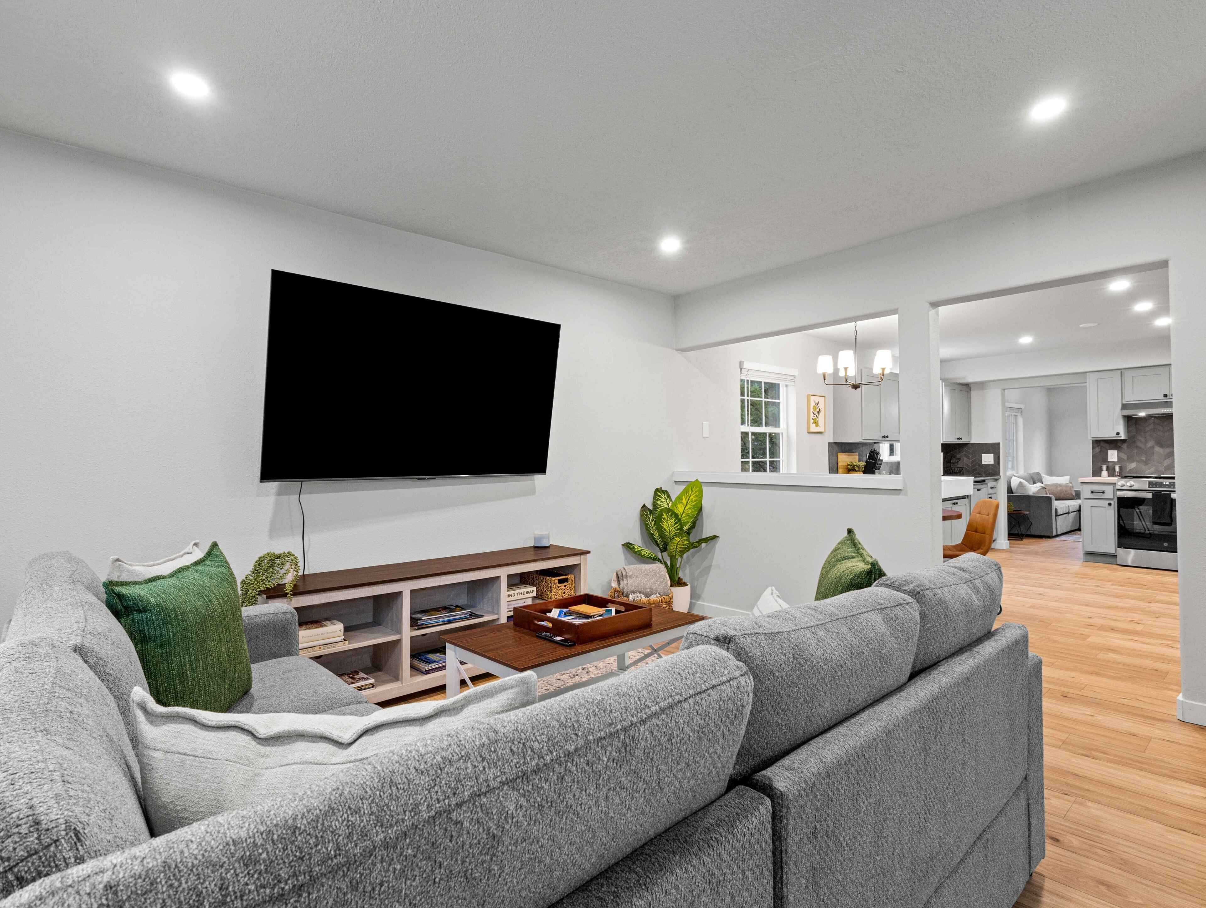 A modern living room with a large flat-screen TV, a grey sofa, a wooden coffee table, and a small bookshelf.
