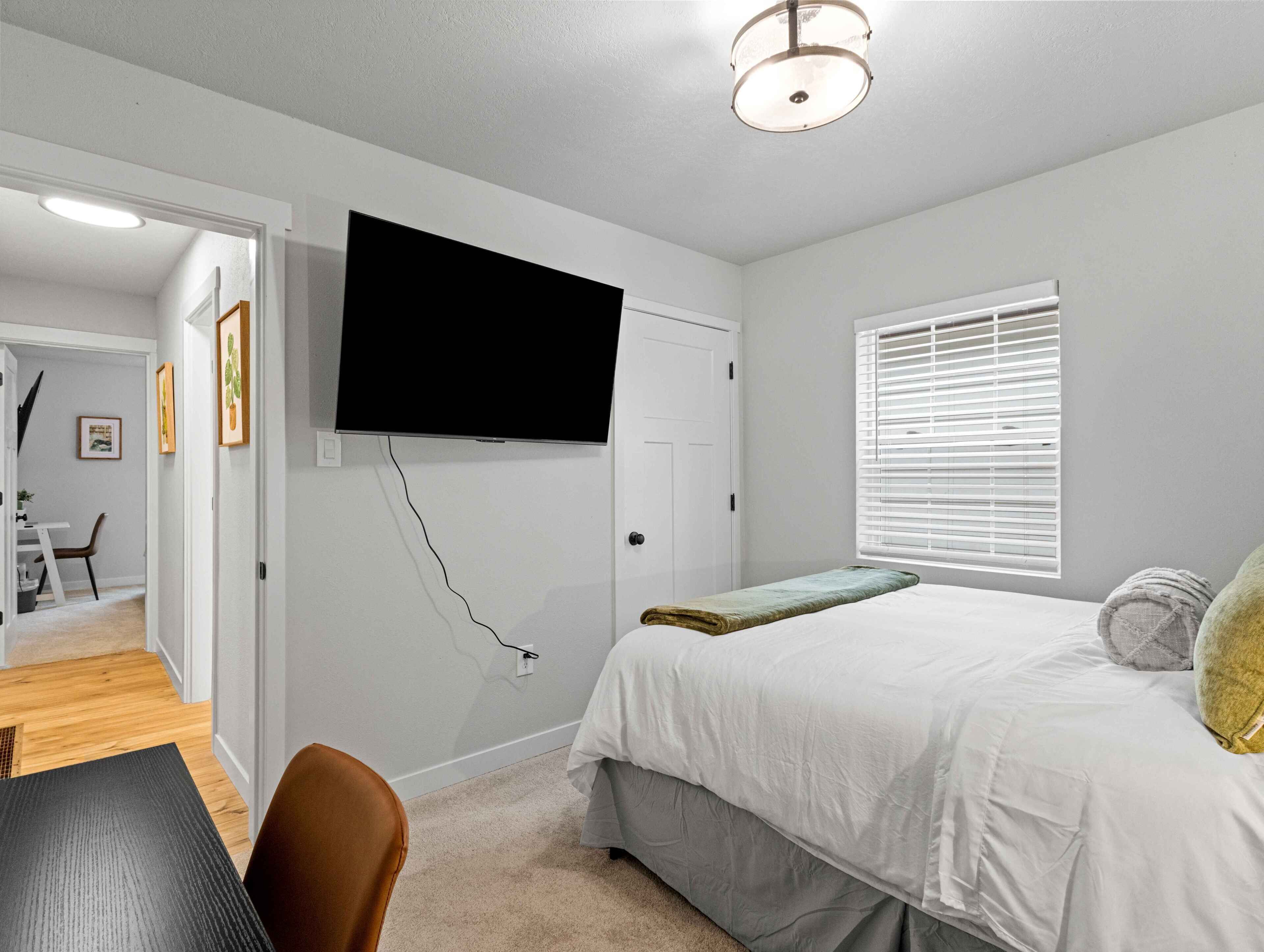A bedroom with a bed, a television mounted on the wall, a window with blinds, and a door leading to another room.