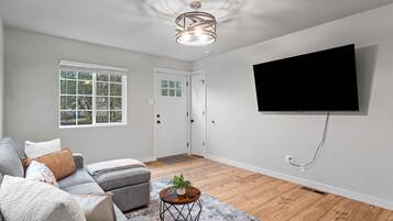 Un salón con un sofá gris, un televisor montado en la pared y una ventana con persianas.