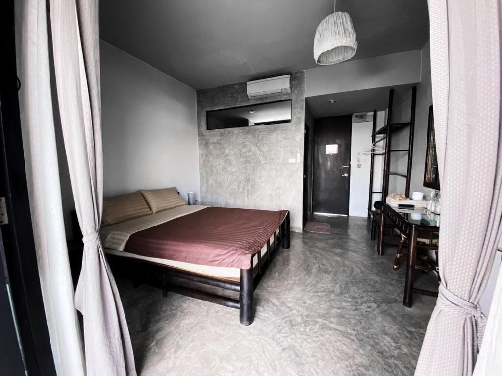 A bedroom with a bed, a desk, a chair, an air conditioning unit, and a hanging light fixture.