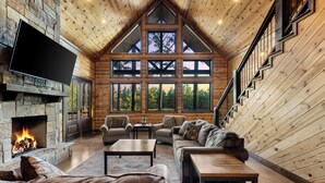 A log cabin interior with a stone fireplace, wooden walls, a large window, and a staircase. - 4 King Suits ~ Deck ~ Playground ~ Hot Tub ~ EV (Broken Bow)