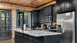 A modern kitchen with dark cabinetry, a stainless steel refrigerator, and a granite countertop. - 4 King Suits ~ Deck ~ Playground ~ Hot Tub ~ EV (Broken Bow)