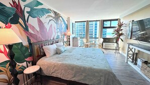 A hotel room with a large bed, a TV, a balcony with a view, and a tropical-themed wall mural. - Tropical Tranquility - Oceanside Retreat w/ Pool (Daytona Beach)