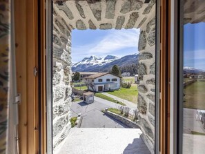Ein Fenster im Steinrahmen mit Blick auf ein Haus und schneebedeckte Berge.