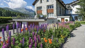 Un edificio de piedra con balcón, rodeado de flores coloridas y montañas al fondo.