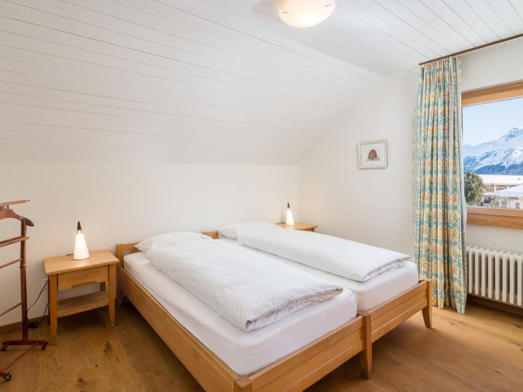 A room with two beds, a wooden floor, a radiator, a window with curtains, and a picture on the wall.