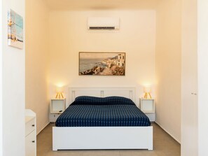 Ein Schlafzimmer mit einem weißen Bett, zwei Nachttischen mit Lampen, einem Bild an der Wand und einer Klimaanlage über dem Bett.
