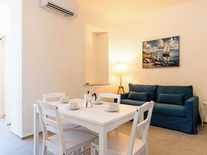 Ein minimalistisches Wohnzimmer mit Essbereich, mit einem weißen Tisch, gedeckten Tassen und einer Kaffeemaschine, einem türkisfarbenen Sofa und einem Gemälde einer Küstenlandschaft.