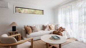 Un salon moderne avec un canapé, une table basse et une lampe. Une fenêtre est agrémentée de rideaux légers et offre une vue sur la verdure à l’extérieur.