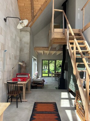 A spacious room with a wooden staircase, a desk with a lamp, and a large window offering a view of the outdoors.