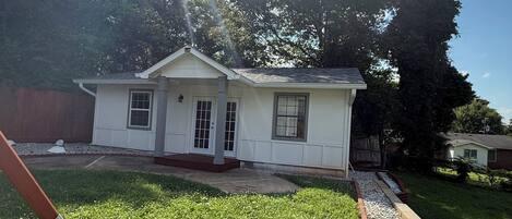 A single-story house with a front porch, a small front yard with grass and a gravel path, and a wooden fence in the background.