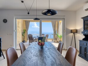 Un espace repas avec une longue table, des chaises, un saladier rempli de fruits et une vue sur la mer grâce à de grandes fenêtres.
