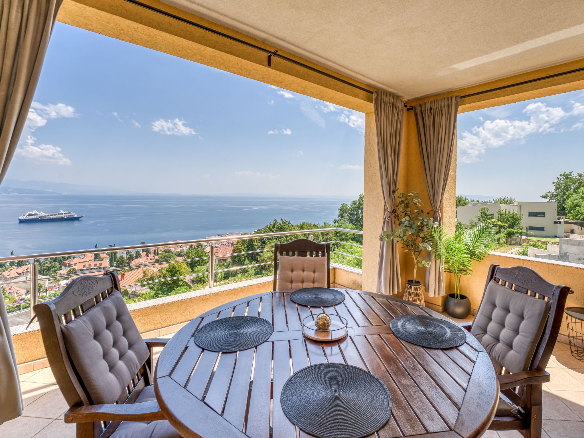 A round wooden table with four chairs, a potted plant, and a view of the sea.