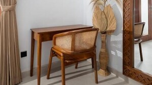 A room with a wooden table, a wicker chair, a tall vase with dried plants, a large mirror, and a beige curtain. - Casa Vara Uluwatu (Badung)