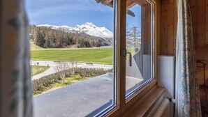 Una habitació amb una finestra gran que dona a una muntanya nevada, un camí i un camp verd.
