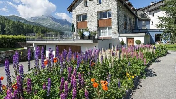 Un edifici de pedra amb un balcó, envoltat de flors colorides i muntanyes al fons.