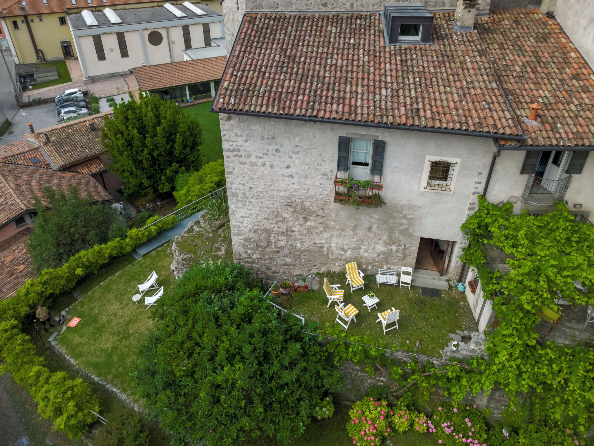 Un bâtiment en pierre avec un toit en tuiles, un petit jardin avec des chaises et un balcon visible, agrémenté de plantes.