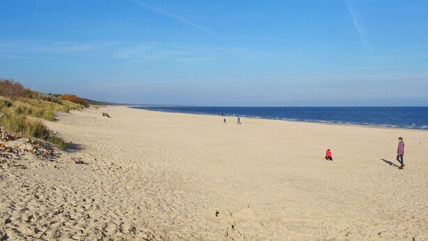 Ein weiter Sandstrand bei blauem Himmel, einige Personen laufen am Strand entlang.