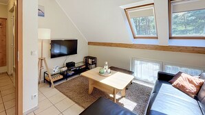 Ein modernes Loft-Zimmer mit schrägem Dach, einem Wand-Fernseher, einem Holztisch, einer Couch und einem kleinen Bücherregal.