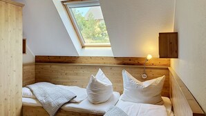 Ein kleiner, gemütlicher Raum mit schrägem Dach, einem Oberlicht, einem hölzernen Bett mit weißer Bettwäsche und einer Wandlampe.