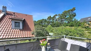 Ein Balkon mit Korbstühlen, einem Tisch mit Buch und Gläsern, einer Vase mit Blumen und Blick auf ein Gebäude mit rotem Dach sowie viel Grün.