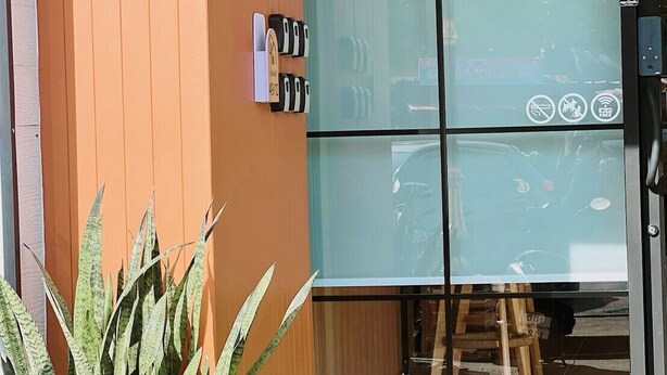 Entrada moderna de un edificio con una planta en maceta, un par de zapatos sobre césped artificial y una puerta de cristal con cerradura electrónica.
