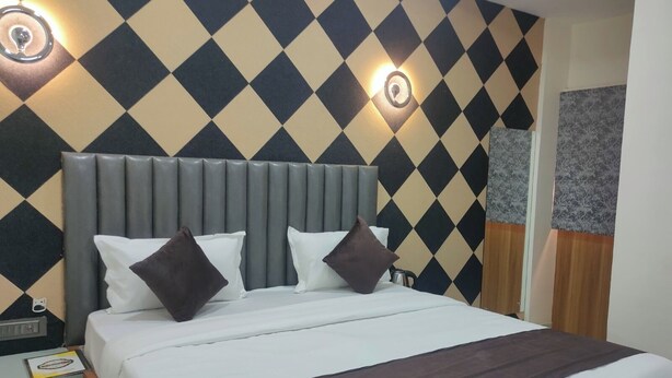 Una habitación de hotel con un estampado de cuadros en la pared, un cabecero tapizado en gris, una cama blanca con almohadas marrones y una mesita de noche con una lámpara.