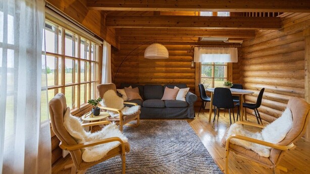 En stuga byggd av timmer med en soffa, stolar, ett matbord och ett stort fönster.