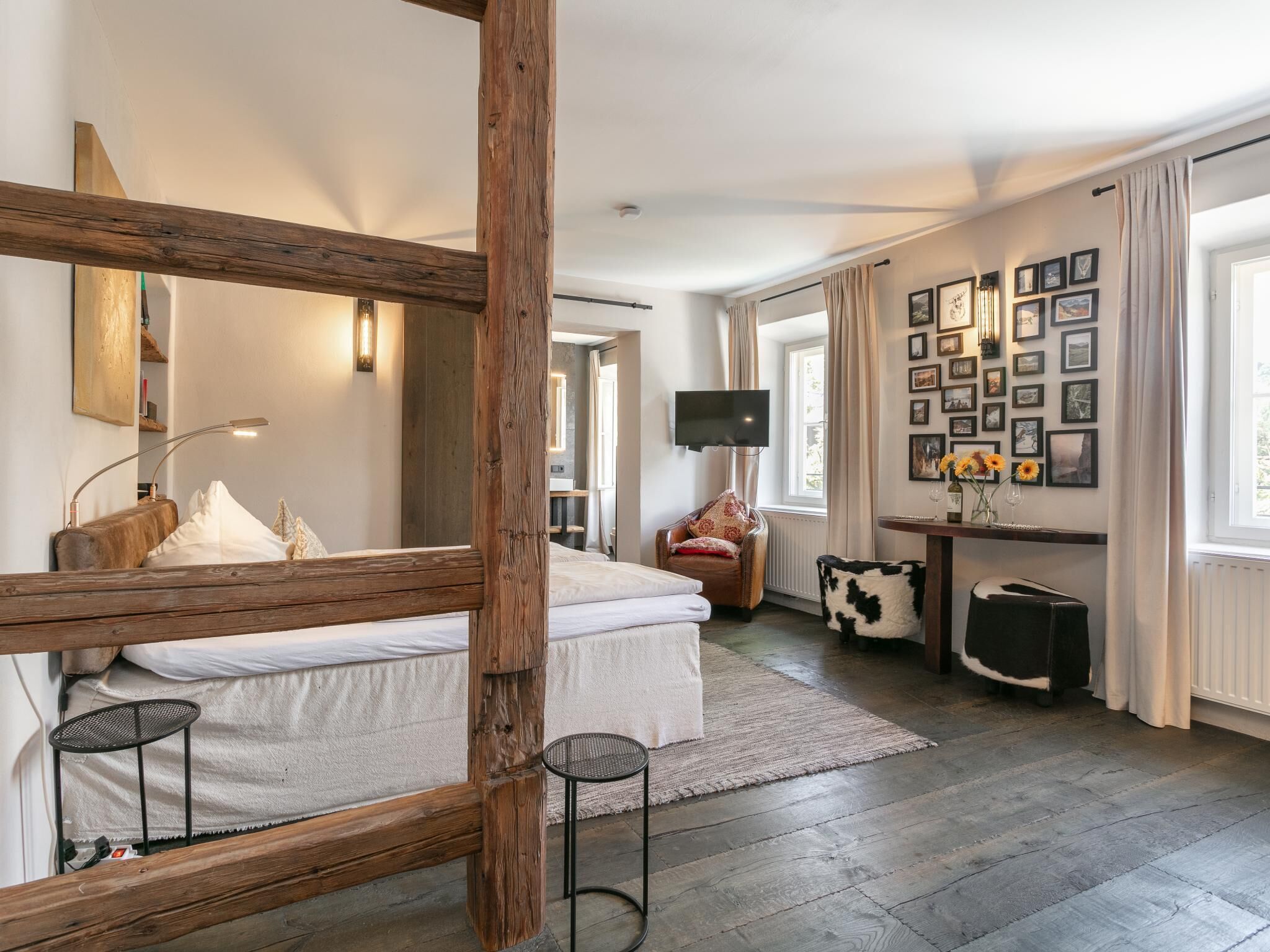 Quarto de hotel moderno com estrutura de cama de madeira, um espelho grande, televisão, área de estar com uma cadeira e uma mesinha, e uma parede decorada com quadros.