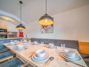 Ein moderner Essbereich mit einem langen Tisch, gedeckt für vier Personen, Pendelleuchten an der Decke und einer Küche im Hintergrund.