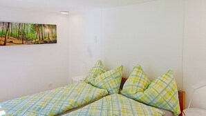 Ein Zimmer mit einem Bett, das eine karierte Tagesdecke und passende Kissen, eine weiße Wand und ein Waldgemälde zeigt.