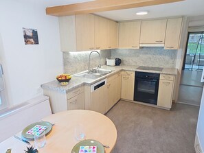 A compact kitchen with a sink, stove, and dishwasher. There is a dining table with plates and a bowl of fruit. - Ferienwohnung ENZian by Interhome (Lungern)