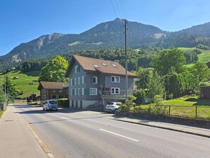 Un bâtiment à deux étages, doté d’un toit en pente, situé à l’angle d’une rue, avec une chaîne de montagnes en arrière-plan.