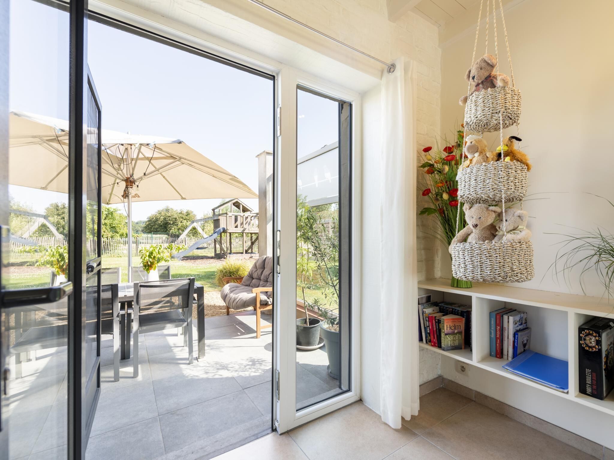 Un interior moderno con un portón corredizo de vidrio que da a un patio, una estantería con libros y una cesta colgante con peluches.