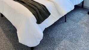 Ein ordentlich bezogenes Bett mit weißen Leinen und einer dunkelgrünen Tagesdecke, eine Nachttischlampe und ein an der Wand hängendes Kunstwerk.