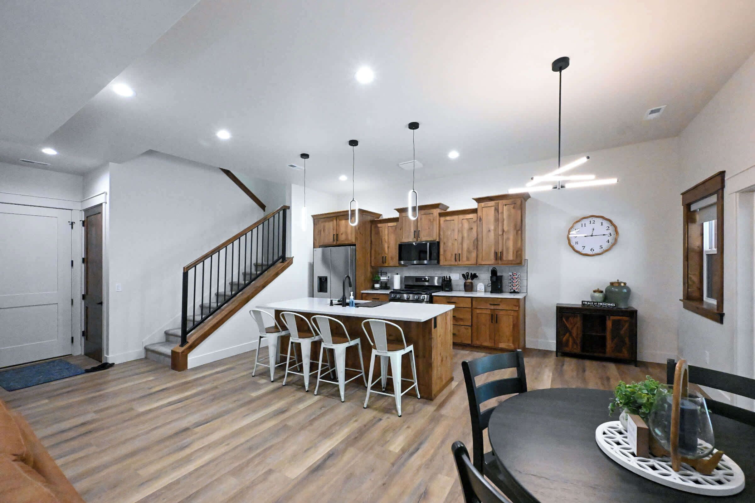 A modern kitchen with wooden cabinets, a central island with bar stools, a dining table with chairs, and a staircase.