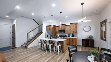 A modern kitchen with wooden cabinets, a central island with bar stools, a dining table with chairs, and a staircase.