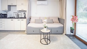 Ett modernt rum med en bäddsoffa, ett litet runt bord, en krukväxt och ett fönster med persienner.