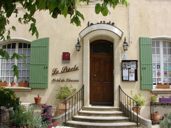 A hotel entrance with a wooden door, green shutters, and a sign reading 'Le Pêche Hotel de Charme.'