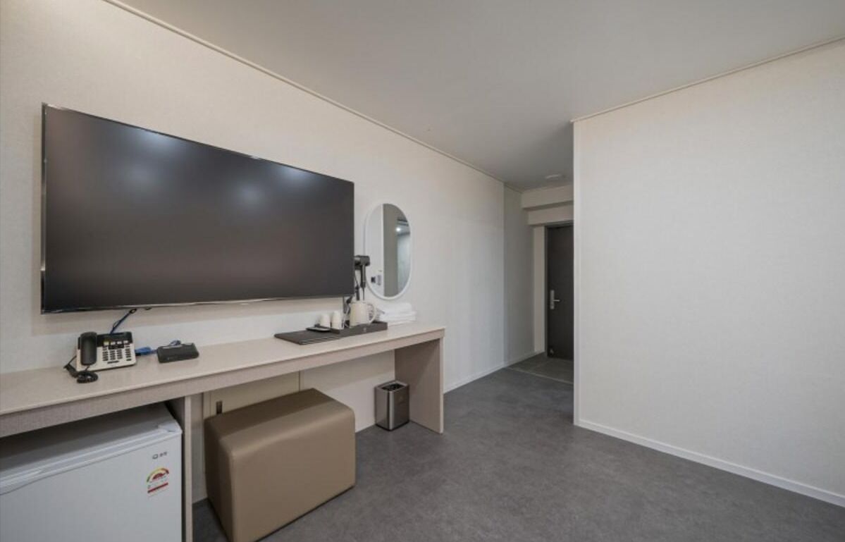 Habitación de hotel con televisor de pantalla plana grande montado en la pared, un escritorio pequeño con teléfono, un espejo y un cubo de basura.