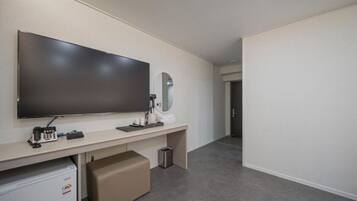 Habitación de hotel con televisor de pantalla plana grande montado en la pared, un escritorio pequeño con teléfono, un espejo y un cubo de basura.